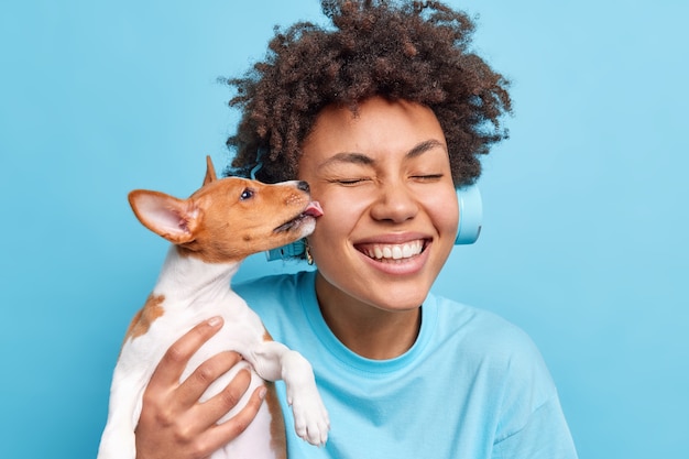 People animals relationship concept. Happy positive curly African American woman poses with pet going to have walk outdoor together. Small dog licks owner in cheek expresses love for being cared