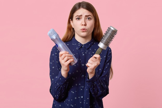 Free photo people, beauty, grooming concept. displeased young woman pouts lips dressed in stylish shirt, poses over rosy wall holds hairspray and comb, comes to hairdresser for making hairstyle.