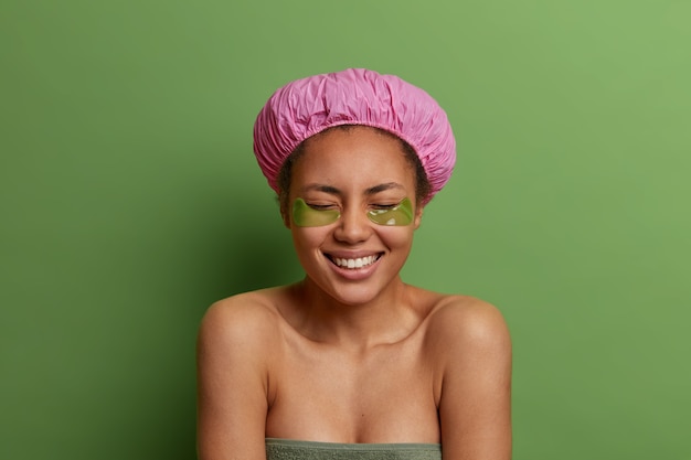人、美容、衛生のコンセプト。うれしそうなアフリカ系アメリカ人の女性は、バスキャップを着用し、タオルで包み、シャワーを浴びた後、眼帯の下に適用し、肌を気にし、緑の壁に対してポーズをとります