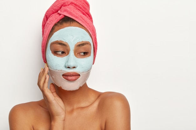 People, beauty, self care concept. Thoughtful dark skinned woman touches cheek, applies clay mask and collagen patch around lips, wears towel on head