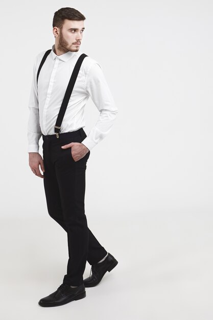 People, beauty, style, fashion, men's wear and accessories. Vertical sideways picture of fashionable young unshaven male modeling in studio, looking back, holding hand in pocket of stylish trousers