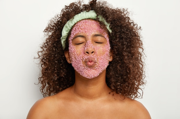 사람, 미용 치료, 건강한 피부 개념. 곱슬 머리를 가진 사랑스러운 아프리카 계 미국인 여성은 입술을 접고 눈을 감고 얼굴이 회춘과 여드름 제거를 위해 장밋빛 소금 알갱이로 덮여 있습니다.