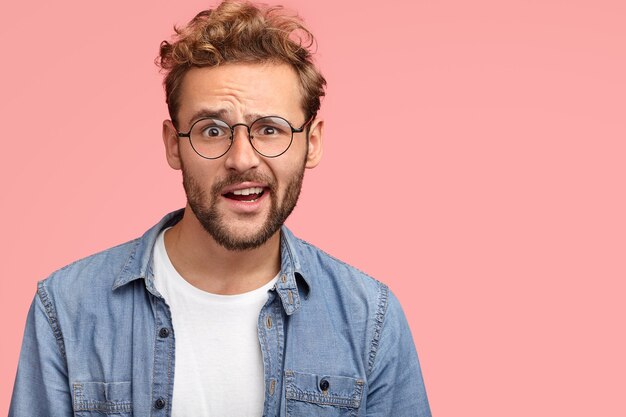 People and bewilderment concept. Puzzled unshaven Caucasian male raises eyebrows surprisingly, being impressed by something, wears white t-shirt and denim shirt, poses against pink wall