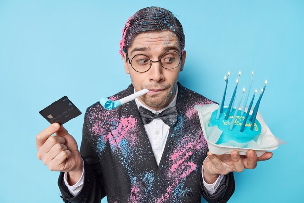 People and birthday celebration concept Man with trendy hairstyle blows party horn holds delicious cake with burning candles and credit card wears festive outfit isolated over blue background