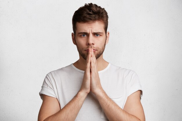 People and body language concept. Handsome serious young male keeps palms pressed together, believes in good luck before important event in life,