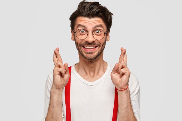 People, body language and expectation concept. Glad Caucasian young male with stubble, keeps fingers crossed, wears white shirt with red braces, has happy expression, isolated over wall