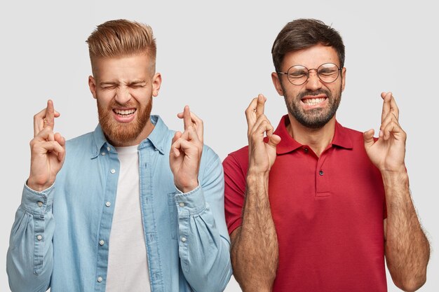 People, body language and wish concept. Dreamy bearded young male hipsters clench teeth and cross fingers, stand closely, hope dreams come true, have specific appearance, isolated over white wall