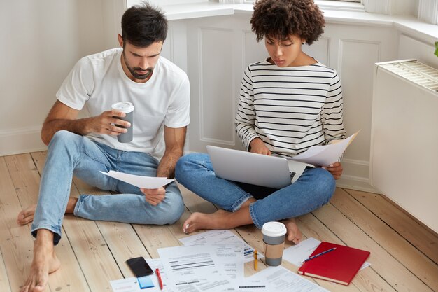 People, business and work concept. Woman and man coworkers study documentation and think about productive strategy to raise profits, pose on wooden floor with takeaway coffee, have serious look