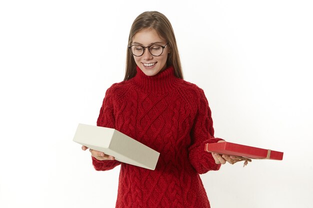 Люди, праздник, радость и счастье. Радостная симпатичная девушка в очках и теплом пуловере стоит с открытой коробкой, смотрит внутрь с заинтересованным любопытным выражением лица, получает подарок на день рождения