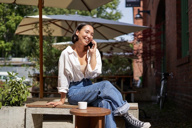 Free photo people and communication stylish asian girl sits in outdoor cafe with cup of coffee and smartphone