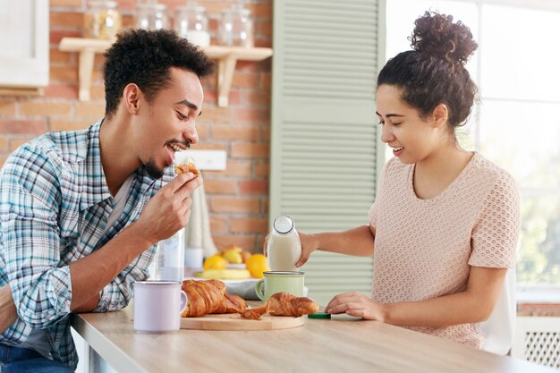 人、料理、試飲のコンセプト。家族カップルが居心地の良いキッチンで昼食をとる：黒い肌のひげを生やした男がおいしい甘いクロワッサンを食べる