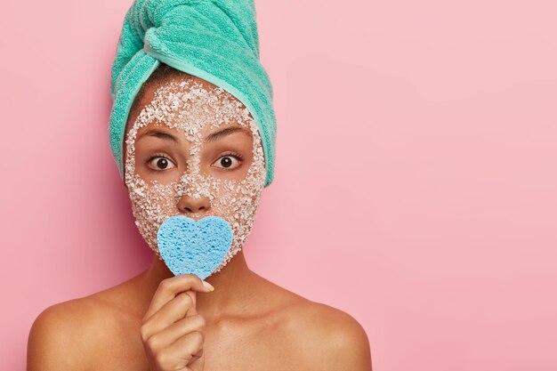 People, cosmetology and wellness concept. Indoor shot of surprised young female model stands naked indoor, covers mouth with blue heart shaped cosmetic sponge, wears facial beauty mask for purity
