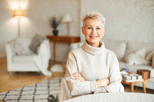 People, coziness, domesticity and season concept. Charming beautiful retired woman spending leisure time indoors at home with confident smile, keeping arms folded on her chest