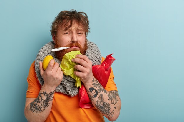 People, disease, healthcare and influenza concept. Dejected red haired bearded guy has high body temperature, suffers from bad feeling, holds tissue, hot bottle and lemon, cures illness at home