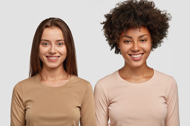 People, diversity and friendship concept. Two happy mixed race beautiful females smile positively, dressed in casual jackets, stand next to each other, isolated over white wall.