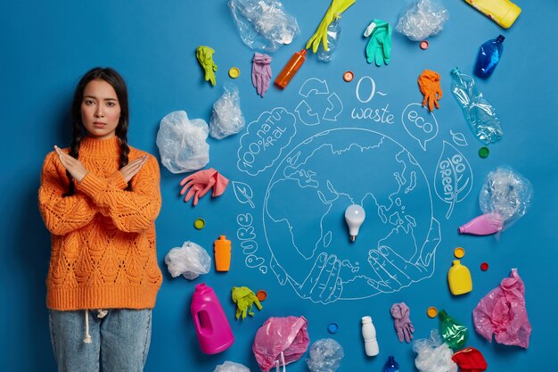 People, ecology, prohibition, refusal concept. serious Asian girl keeps arms crossed over chest, says no to plastic, being eco friendly, stands against blue wall