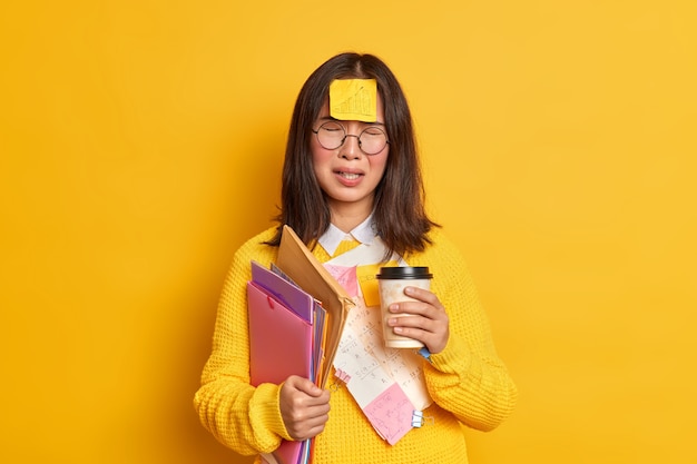 People and education concept. Upset Asian female student feels tired of exam preparation drinks takeaway coffee makes memo stickers has bad mood cannot remember everything holds folders with papers