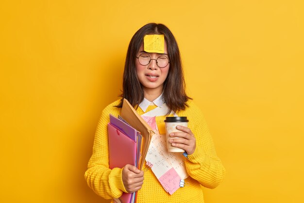 Концепция людей и образования. Расстроенная азиатская студентка чувствует себя усталой от подготовки к экзамену, выпивает кофе на вынос, делает стикеры-памятки в плохом настроении, не может вспомнить все, держит папки с бумагами