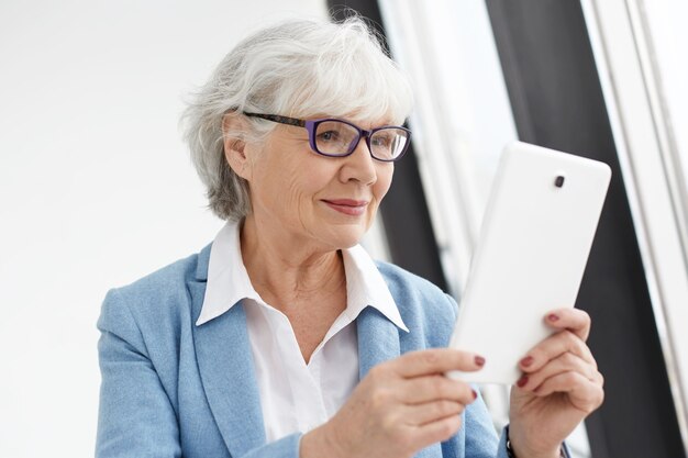 People, electronic gadgets, technology and communication concept. Modern smart mature senior woman entrepreneur in stylish suit and rectangular glasses holding digital tablet, surfing internet