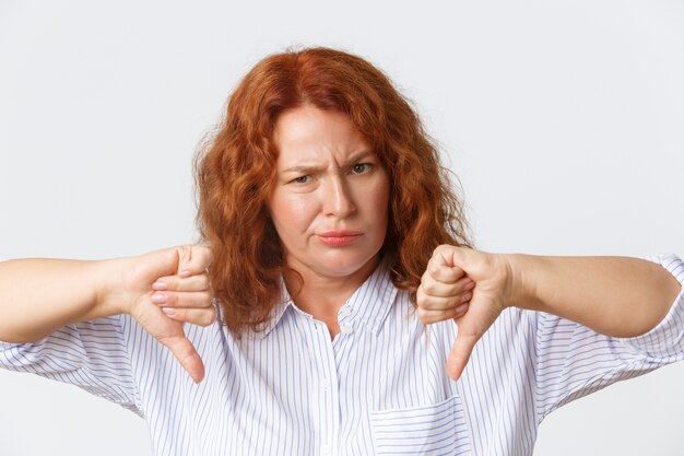 People, emotions and lifestyle concept. Close-up of disappointed and upset, frowning middle-aged redhead woman looking bothered and unamused, showing thumbs-down, dislike and disapproval gesture.