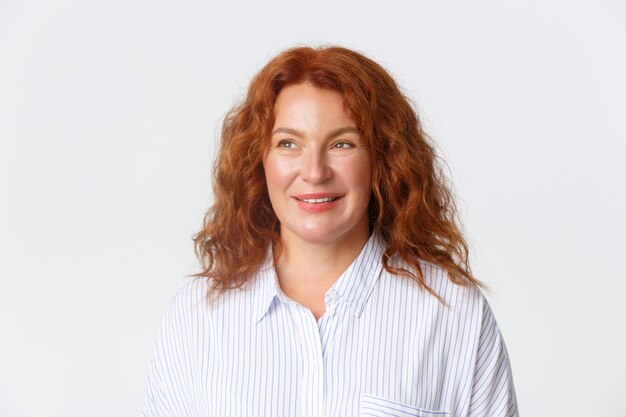 People, emotions and lifestyle concept. Dreamy and happy middle-aged redhead woman in blouse smiling pleased, looking left with hopeful gaze, standing white background daydreaming.