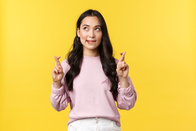 People emotions, lifestyle and fashion concept. Dreamy and hopeful cute asian girl having aspirations, biting lip and looking upper left corner tempting, cross fingers good luck, yellow background.
