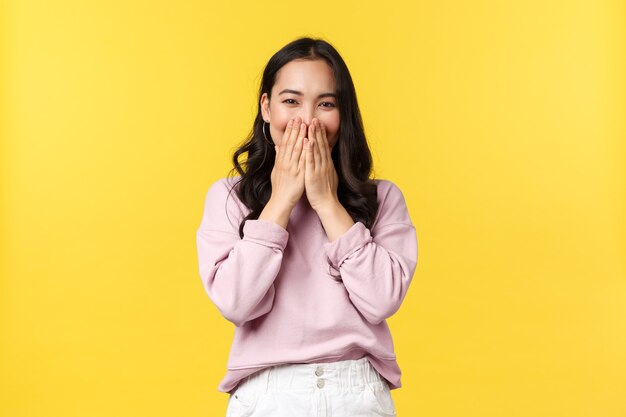 People emotions, lifestyle and fashion concept. Funny and cute korean woman laughing shy, smiling with eyes while cover mouth and giggle silly at camera, standing yellow background.