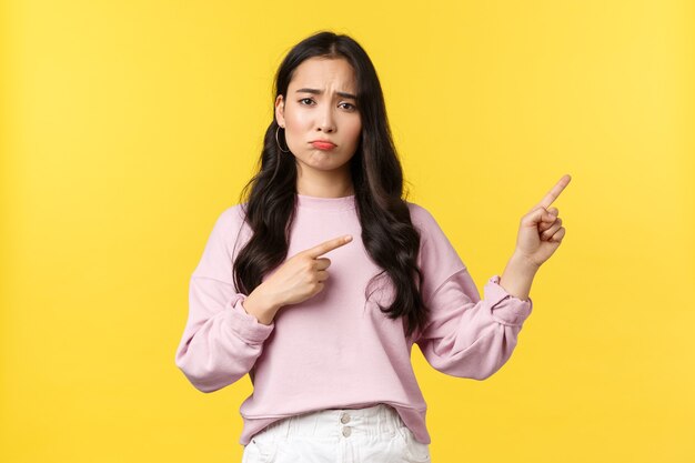 People emotions, lifestyle and fashion concept. Gloomy complaining asian woman pointing fingers right and sulking from regret or disappointment, feeling sad over yellow background.