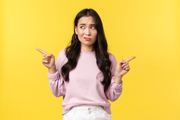 Free photo people emotions, lifestyle and fashion concept. indecisive troubled asian woman having problem with choice, pointing fingers sideways and smirk questioned, looking left, deciding, yellow background.