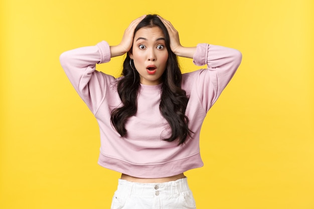 People emotions, lifestyle and fashion concept. Overwhelmed and shocked asian girl grab head and react to concerning big news, saying wow, stare camera astonished, yellow background.