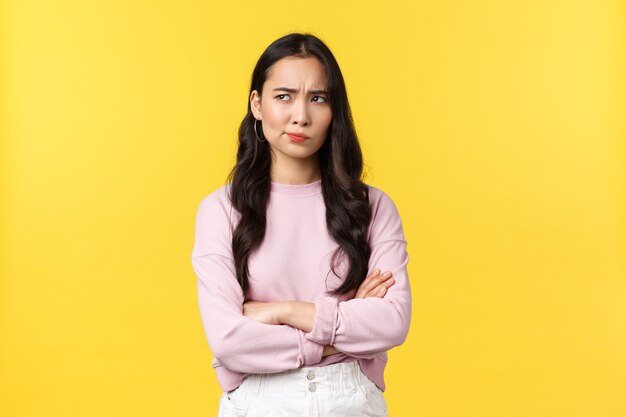 People emotions, lifestyle and fashion concept. Troubled and doubtful asian girlfriend having concerning thoughts, looking upper left corner and grimacing, pondering, yellow background.