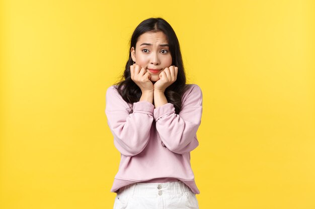 People emotions, lifestyle and fashion concept. Worried thoughtful asian girl holding hands pressed to chin and looking away dreamy, feeling nervous before important interview, yellow background