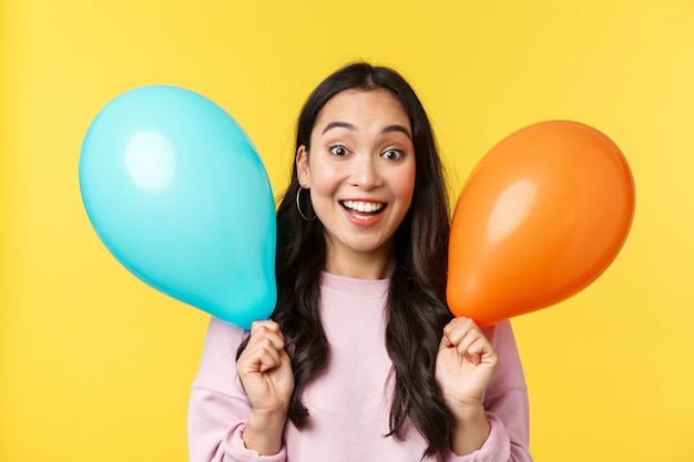 People emotions, lifestyle leisure and beauty concept. Cheerful happy asian girl congratulate with big holiday, holding two balloons and smiling broadly, enjoy party over yellow background.