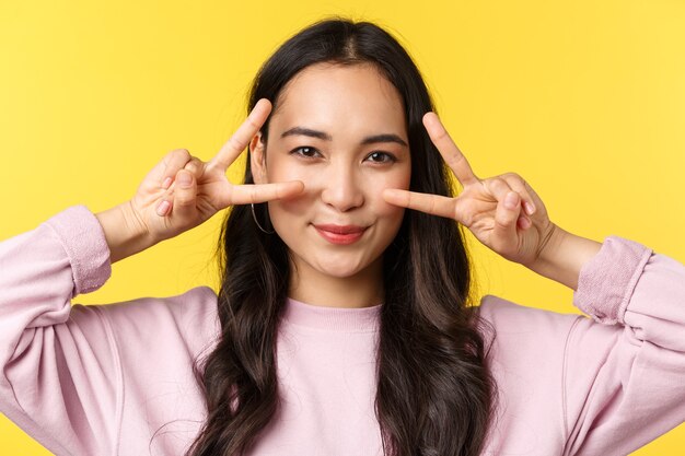 People emotions, lifestyle leisure and beauty concept. Close-up of optimistic kawaii asian girl making peace signs over eyes and smiling, show positive happy expression, yellow background.