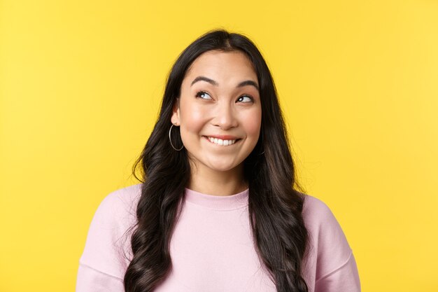 People emotions, lifestyle leisure and beauty concept. Excited and cute silly asian girl cant wait something happen, biting lip from temptation and looking upper left corner happy, yellow background.