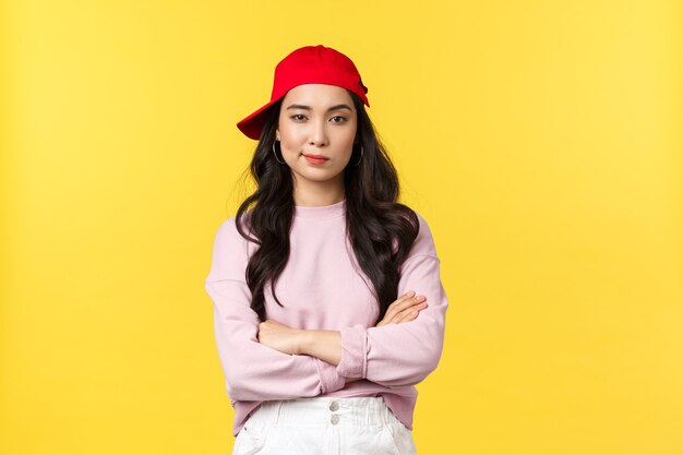 사람들의 감정, 라이프 스타일 여가 및 아름다움 개념. 빨간 모자를 쓴 진지해 보이는 자신감 있는 아시아 여성, 시원하고 건방지게 보이는, 팔짱을 끼고 있는 가슴, 노란색 배경에 서 있는.