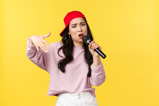 People emotions, lifestyle leisure and beauty concept. Stylish and cool young girl rapper in red cap, singing song and gesturing, performing with microphone, standing yellow background.