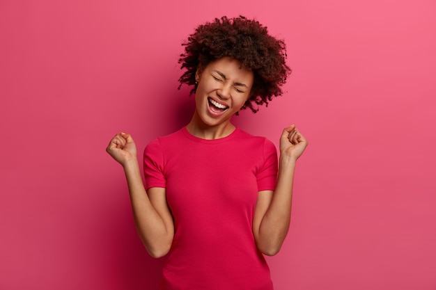 People, emotions, triumph and success concept. Happy Afro American teenage girl celebrates victory, raises fists up, has upbeat mood, dressed in casual clothing, isolated on pink wall.