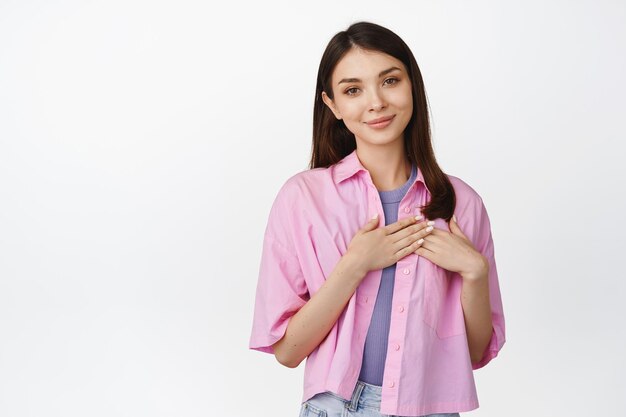 People emotions Young woman holds hands on heart smiles at camera stands against studio background isolated on white