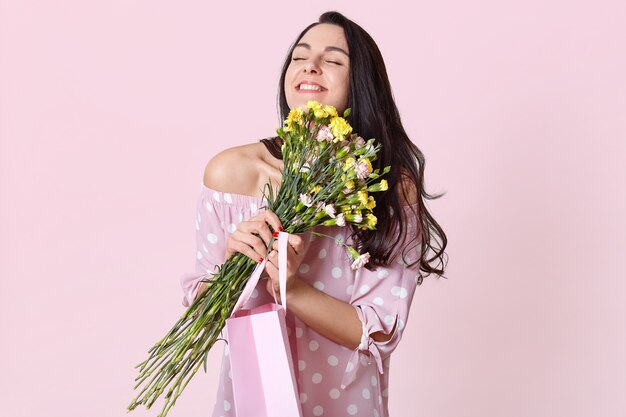 People, enjoyment and happiness concept. Positive dark haired woman with black hair, embraces flowers, carries gift bag, poses on light pink. Female rejoices present on 8 March.