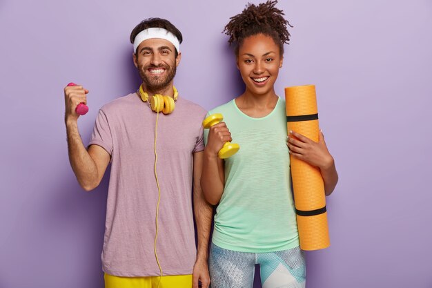 People, exercising and sport concept. Happy Caucasian man and dark skinned woman raise dumbbells, carry fitness mat, have toothy smiles