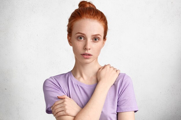 People, facial expressions and emotions concept. Adorable freckled red haired female model has hair knot, dressed in casual clothing