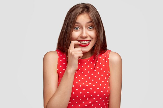 People and fashion concept. Lovely young female giggles positively, keeps finger near mouth, dressed in elegant red dress, has long dark hair, notices something funny, isolated over white wall