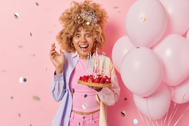 People and festive event concept Cheerful woman looks with glad excited expression dressed in fashionable clothes holds strawberry cake stands near bunch of balloons isolated on pink background