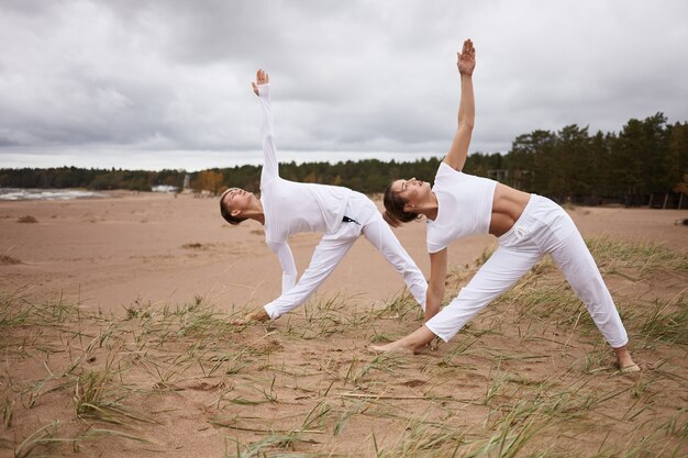 People, fitness, sport, friendship, family and lifestyle concept. Professional female yoga instructor and her teenage son both in white clothes, standing barefooted on sand, doing Utthita Trikonasana