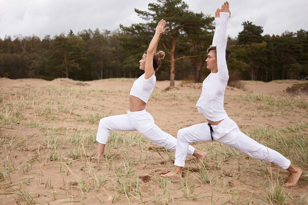 People, fitness,yoga, pilates and active healthy lifestyle concept. Summertime shot of sport barefoot young Caucasian man and woman in white clothes doing Virabhadrasana or Warrior 1 pose outdoors