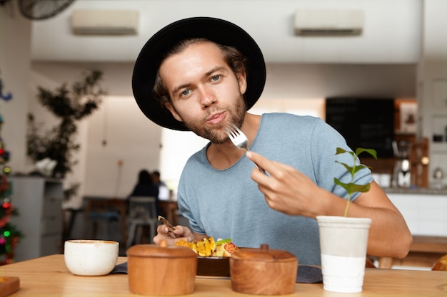 自由摄影人来说,食物和生活方式的概念。室内拍摄漂亮的年轻的学生戴着黑色帽子和蓝色t恤安抚他的饥饿