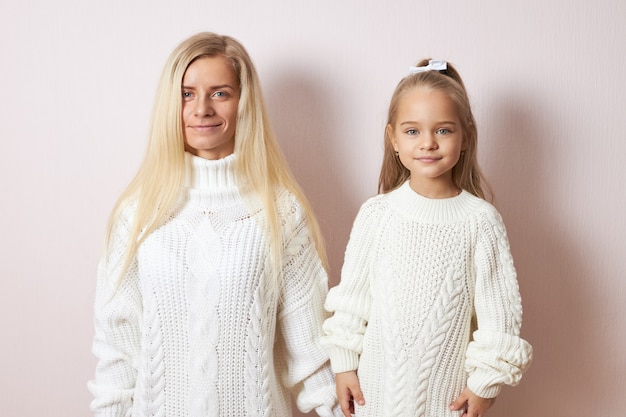 People and generations concept. Isolated shot of attractive young European mother posing  holding hands with beautiful little daughter, both dressed in cozy warm sweaters