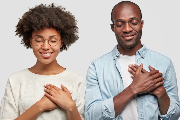 People and gratitude concept. Horizontal shot of pretty young African American female and dark skinned male keep hands on chest, being thankful to people who helped them, have charming smiles