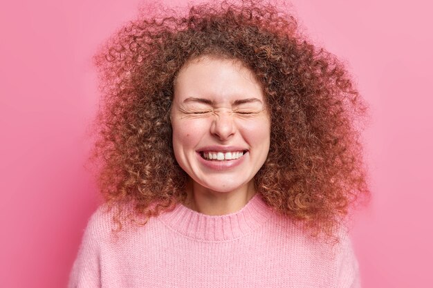 People and happiness concept. Glad curly haired young European woman smiles broadly feels very happy closes eyes squints  dressed in casual jumper isolated over pink  wall.